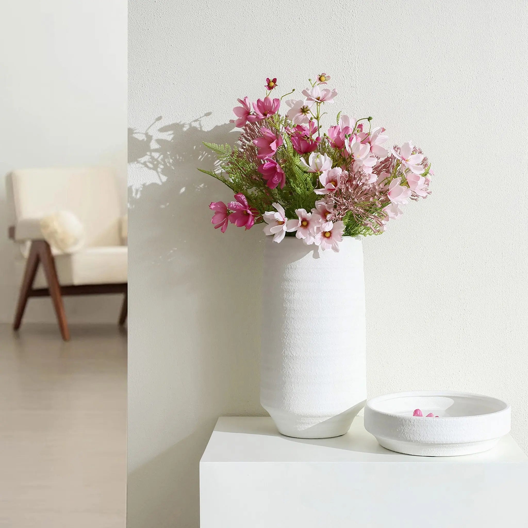 A white terracotta vase with pink and white flowers graces a white table beside an empty bowl. In the background, a beige chair adds warmth to this serene indoor scene.