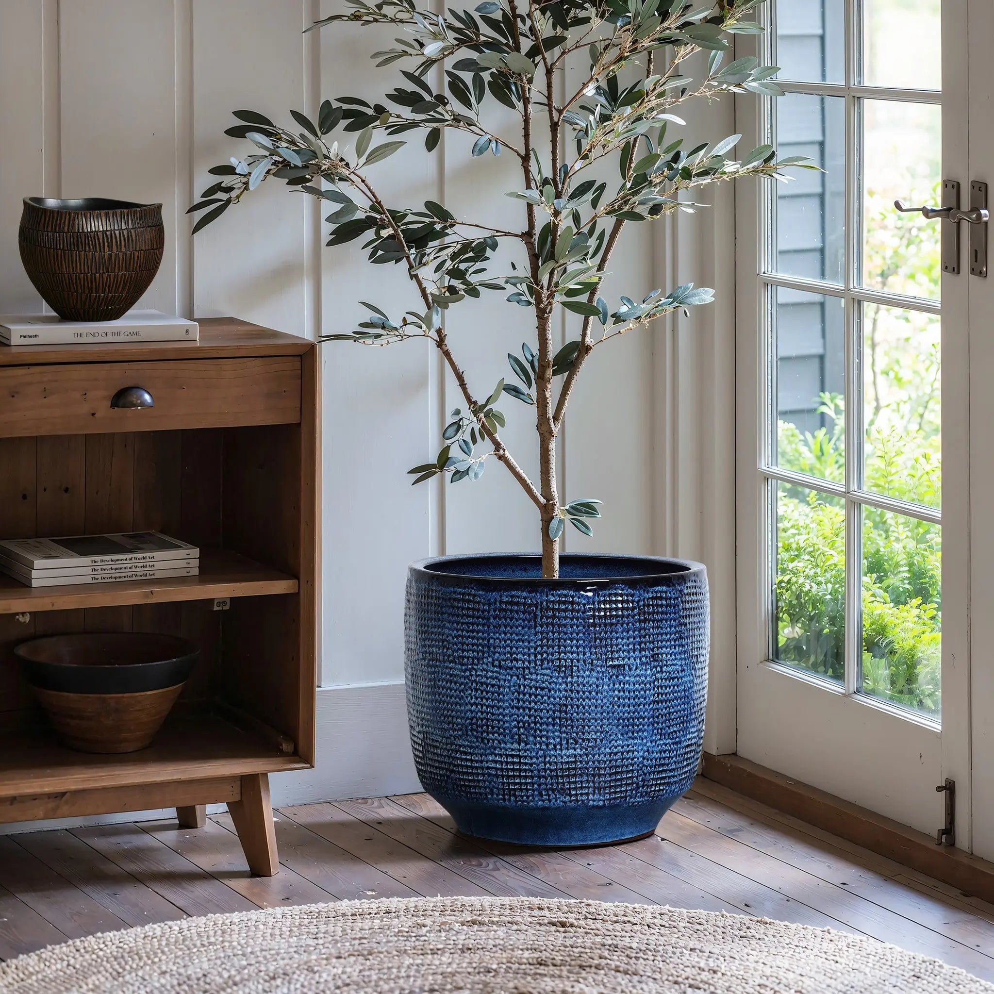 An Ocean blue glazed round ceramic planter (13.98 h) by LuxenHome, holding a tall green plant, sits on a wooden floor near a glass door. To the left is a wooden shelf with books, a decorative vase, and a bowl; while in the foreground, part of a woven rug is visible.