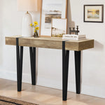 A modern farmhouse console table with black legs and an oak finish features a white lamp, yellow flowers in a vase, two framed pictures, books, and a small candle holder, all set against a crisp white wall.