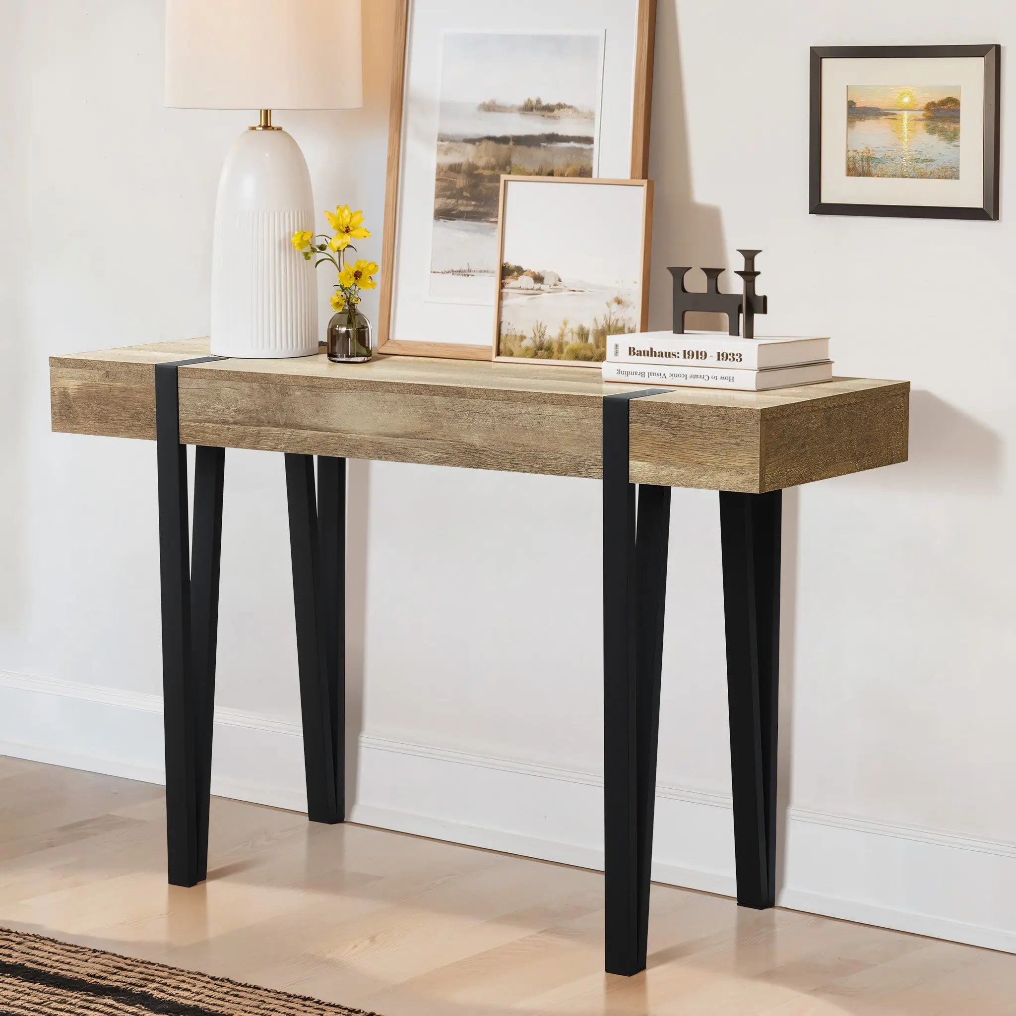 A modern farmhouse console table with black legs and an oak finish features a white lamp, yellow flowers in a vase, two framed pictures, books, and a small candle holder, all set against a crisp white wall.