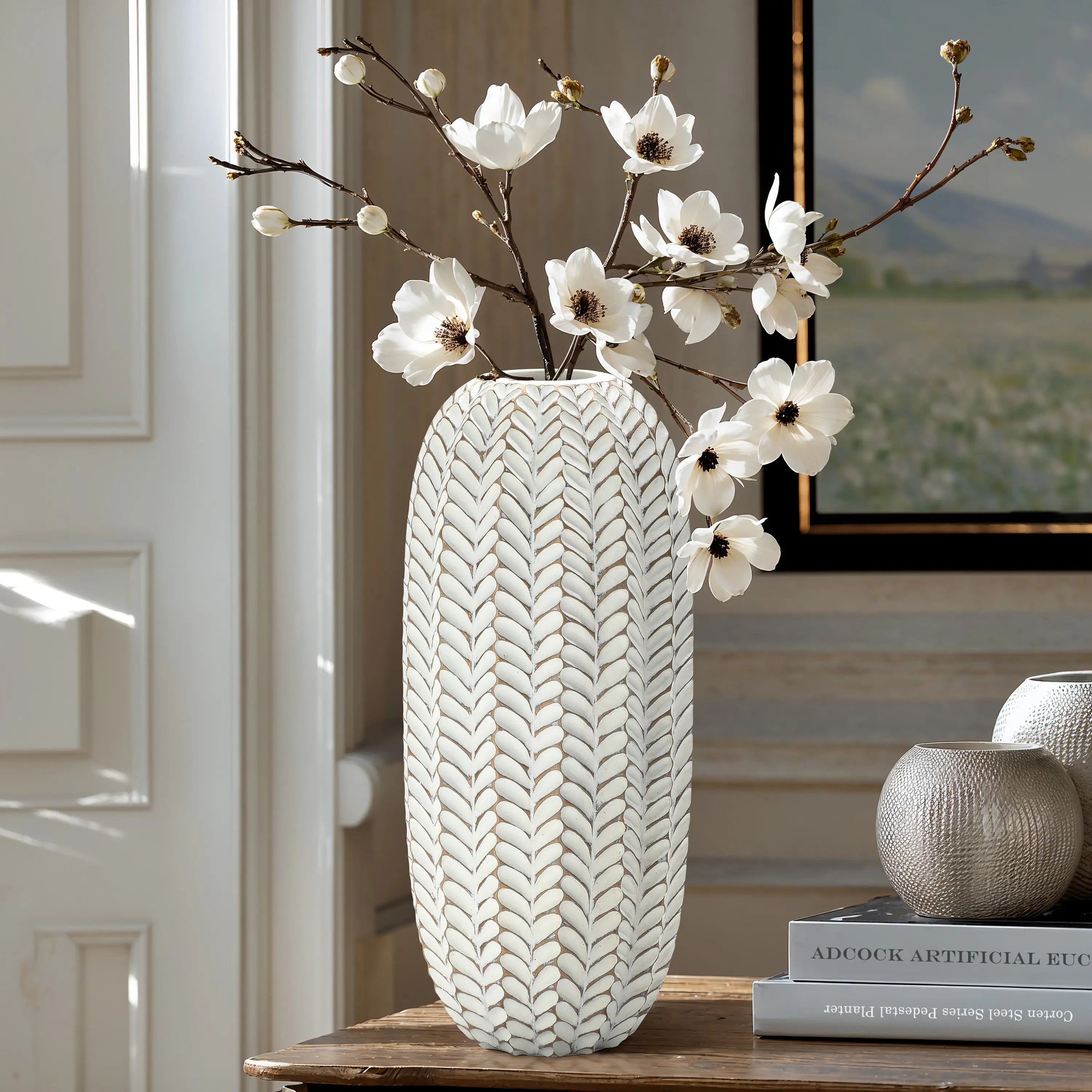 LuxenHomes Modern geometric pattern resin tall vase, 17 h, with an antique effect and white blossoms, stands on a wooden table beside two round vases. Its set against a cozy interior backdrop showcasing a partially visible painting.