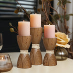 Three lit pink candles sit on handcrafted wooden holders wrapped in twine, placed on a table next to a glass candle holder and a decorative yellow rose, showcasing an elegant rattan design.