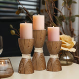 Three lit pink candles sit on handcrafted wooden holders wrapped in twine, placed on a table next to a glass candle holder and a decorative yellow rose, showcasing an elegant rattan design.