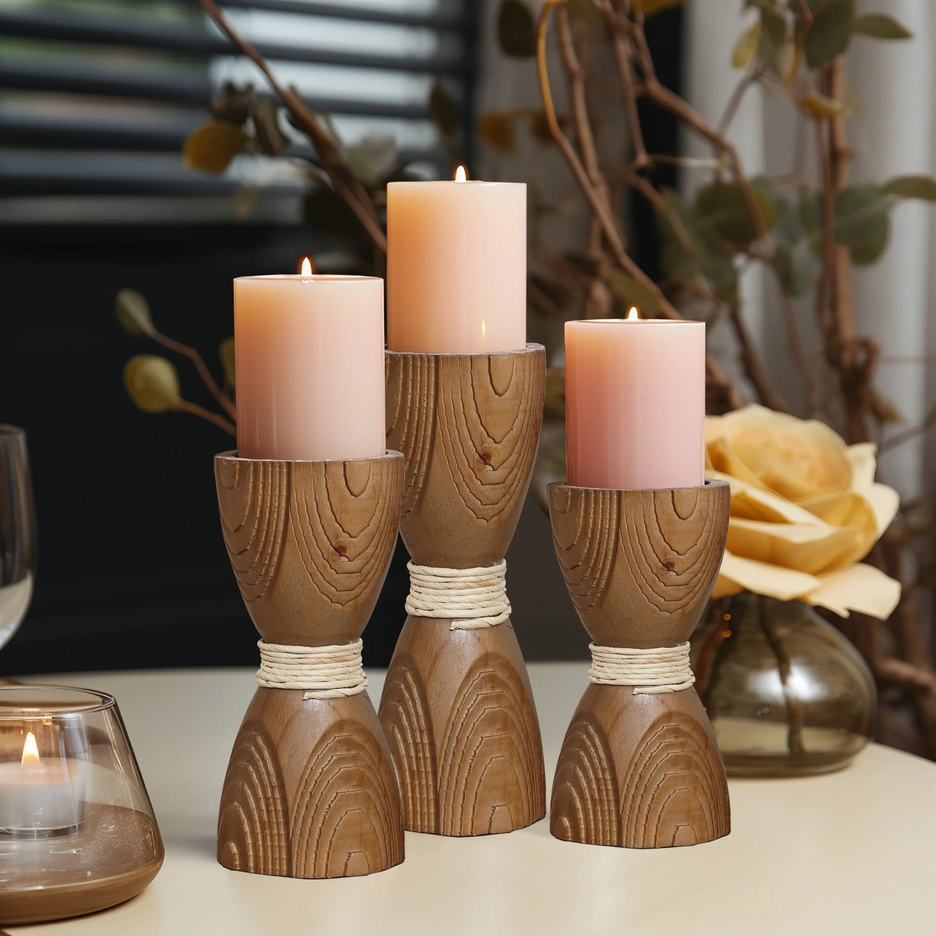Three lit pink candles sit on handcrafted wooden holders wrapped in twine, placed on a table next to a glass candle holder and a decorative yellow rose, showcasing an elegant rattan design.