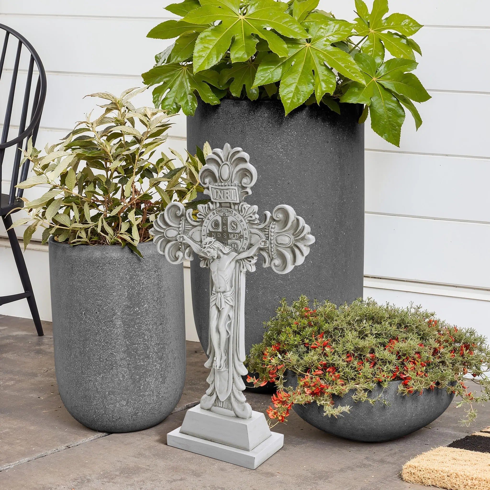 A decorative stone cross stands gracefully beside three potted plants on the patio, with a serene white wall framing the scene. This charming arrangement subtly echoes the Christian faith, inviting peace and reflection in its simplicity.