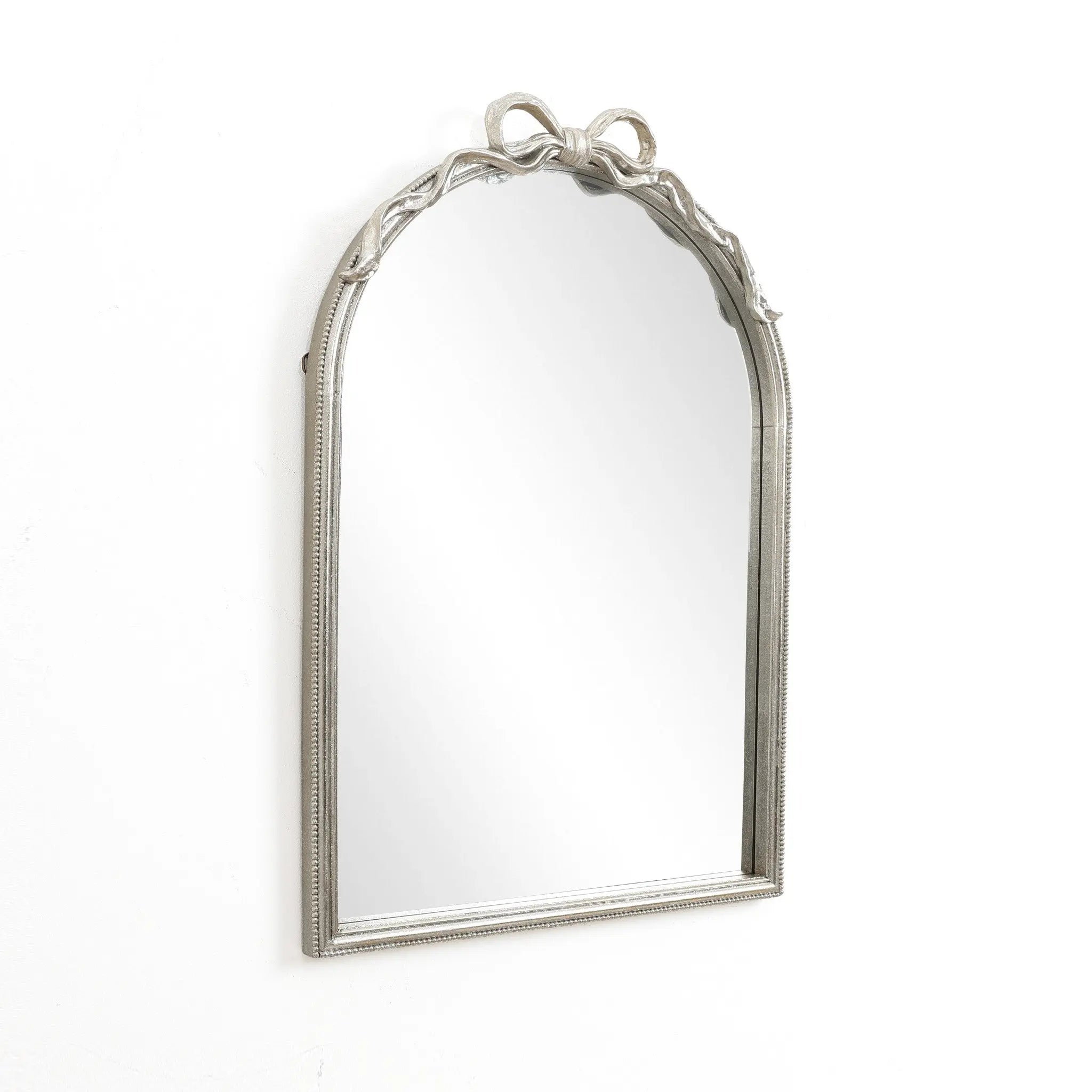 The Ribbon Wall Mirror features an elegant, arched silhouette with a decorative silver frame and a charming bow design atop, embodying a modern romantic style. Set against a plain white background, it effortlessly blends classic elegance with contemporary flair.