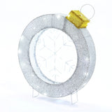 A circular, silver tinsel snowflake wreath with a snowflake design in the center and a golden bow on top stands upright on a small stand, perfect as a festive Christmas decor.
