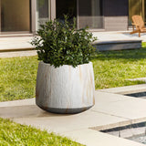 A large white MgO planter with a green leafy plant graces the paved patio, nestled beside a serene water feature. The scene is completed by a charming house and wooden chair in the background, seamlessly blending indoor-outdoor living.