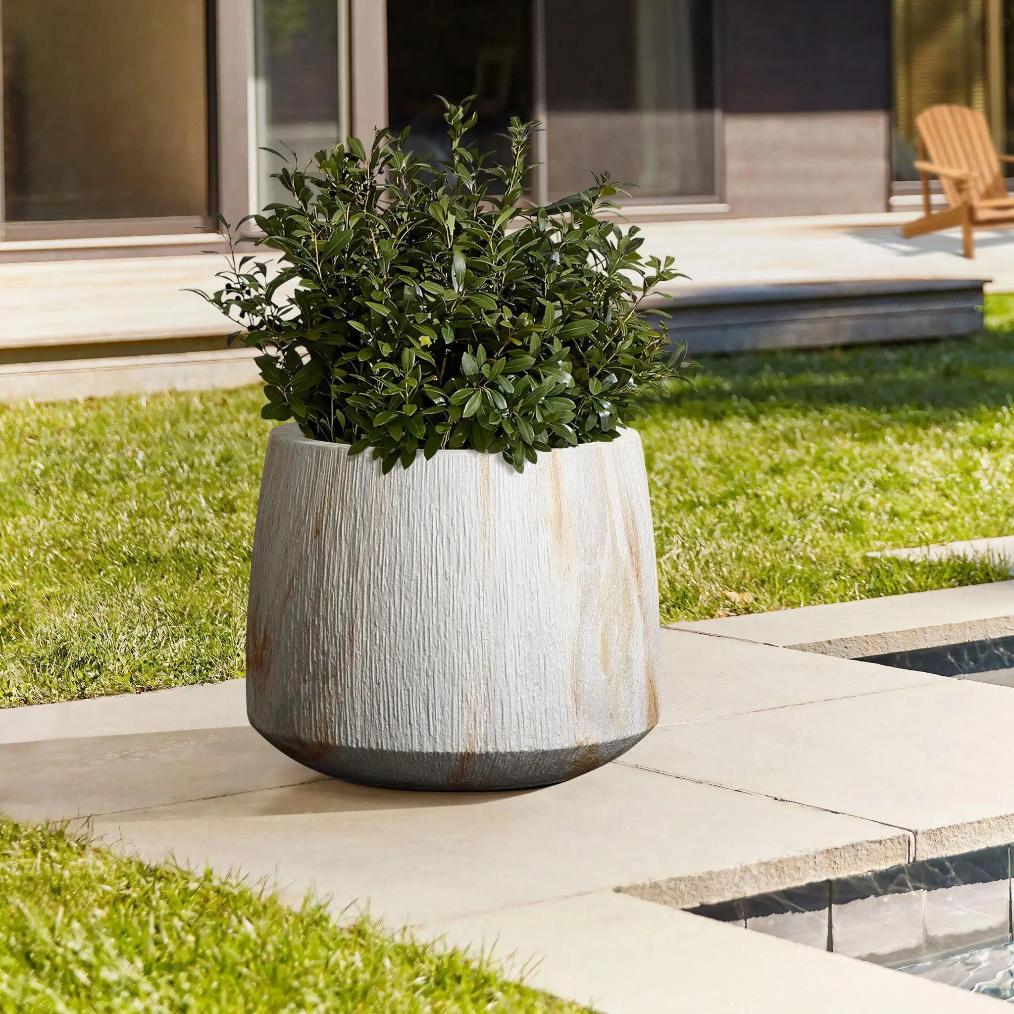 A large white MgO planter with a green leafy plant graces the paved patio, nestled beside a serene water feature. The scene is completed by a charming house and wooden chair in the background, seamlessly blending indoor-outdoor living.
