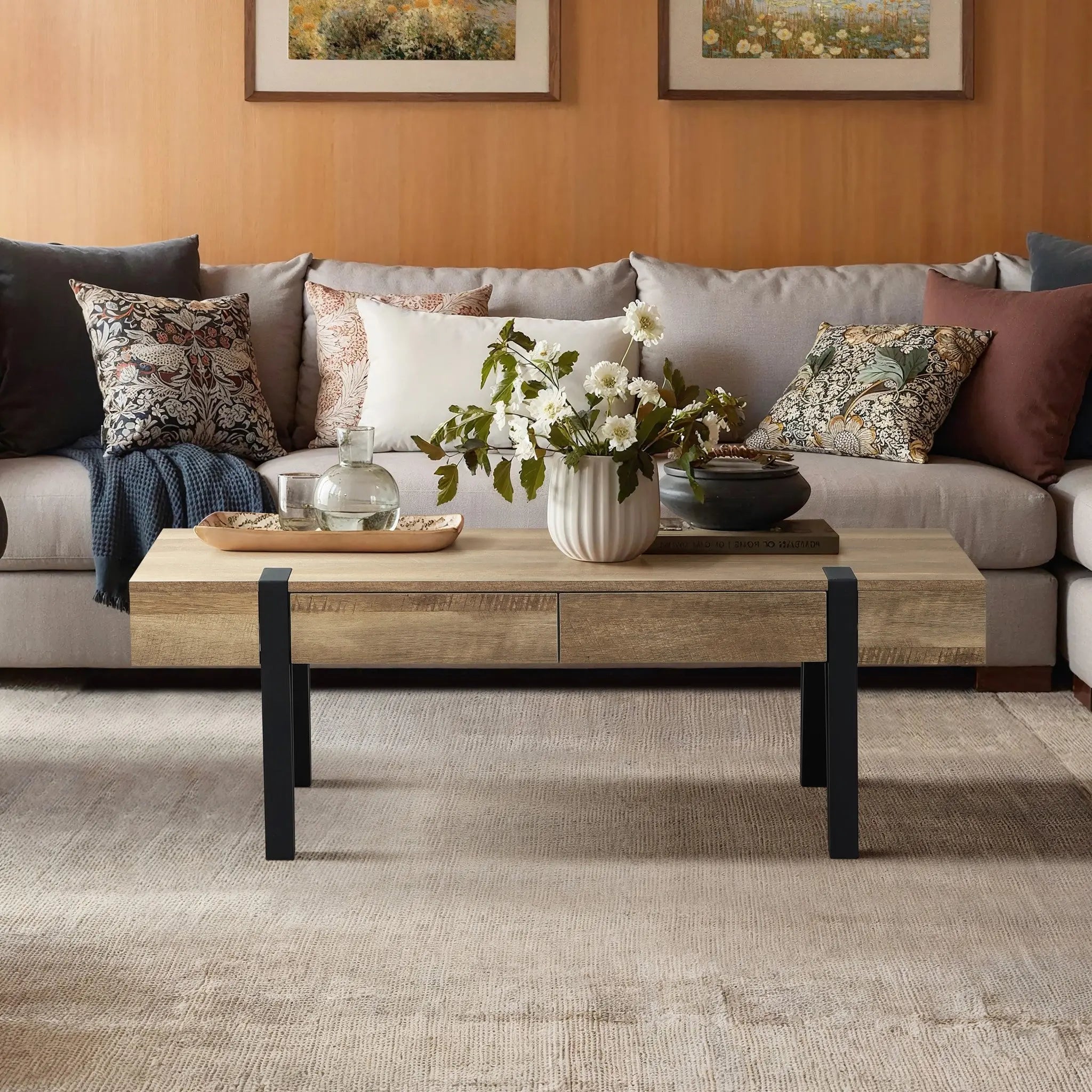 A modern living room with a beige sectional sofa, patterned cushions, and a modern farmhouse coffee table in oak finish wood. A vase of flowers sits on top, while two framed artworks hang on the wall—truly a living room essential.