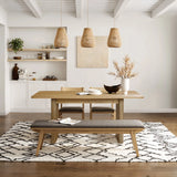 A modern dining area features the stylish LuxenHome Aria wood finish 63 dining table for six, accompanied by two chairs and a cushioned bench on a patterned rug. Three woven pendant lights add warmth, while a shelf with decor and a potted plant enhances the aesthetic against the white wall.