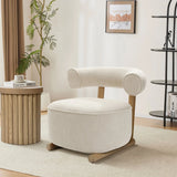 A cozy, contemporary living space showcases the LuxenHome Rio accent chair with a rounded backrest and cream velvet upholstery. It sits on a light rug near a wooden side table with books. A plant, framed artwork, and a decorative shelf with books and decor complete the scene.