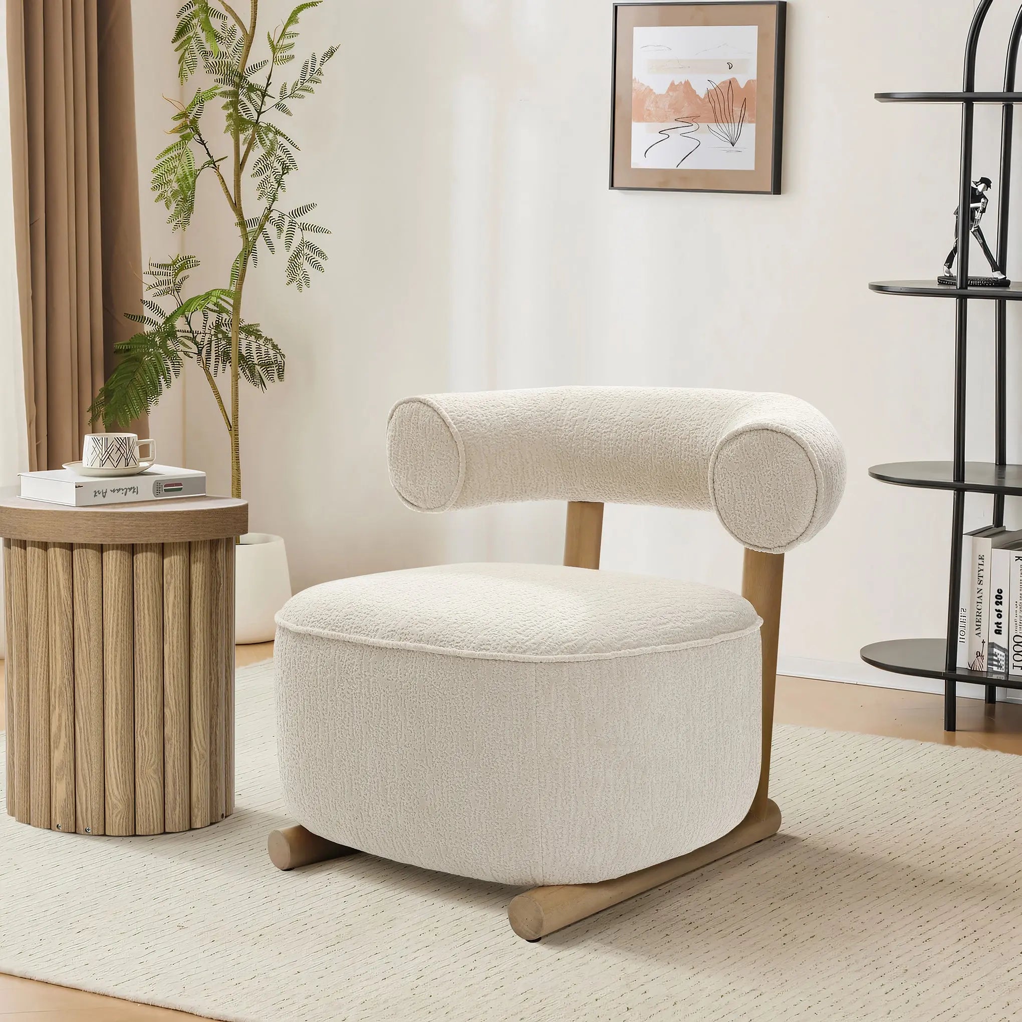 A cozy, contemporary living space showcases the LuxenHome Rio accent chair with a rounded backrest and cream velvet upholstery. It sits on a light rug near a wooden side table with books. A plant, framed artwork, and a decorative shelf with books and decor complete the scene.