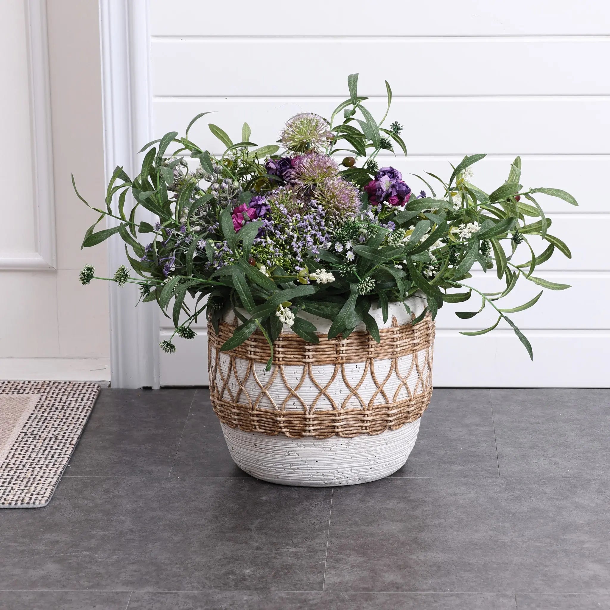 A bohemian-style planter brimming with lush green foliage and vibrant purple flowers sits gracefully on the gray tiled floor, right next to the door.