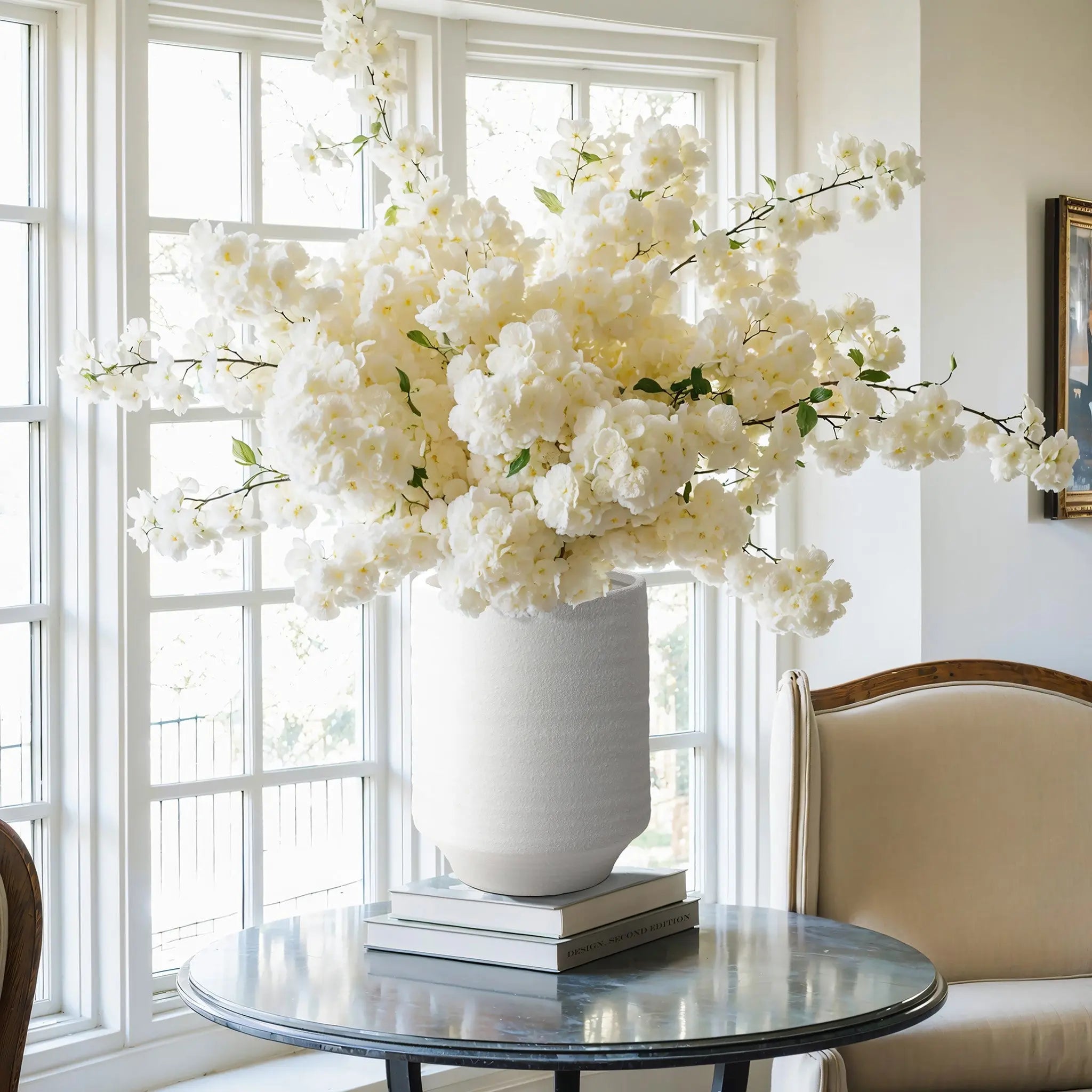 A stunning white terracotta vase, 12 inches tall, is filled with abundant white flowers on a round glass table by the window, complemented by a nearby beige chair.