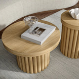 Wooden round coffee tables with fluted bases add a touch of modern design. One table, with an oak finish, has a stack of books and a glass jug on top, while the other features a small white bowl. They sit gracefully on a light-colored textured rug.
