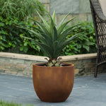 A green agave plant in a large brown contemporary planter sits on a grey stone patio near a stone wall and greenery.
