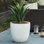 A green, spiky-leaved plant in a weather-resistant planter sits on a gray stone patio near wicker furniture and an outdoor lantern.