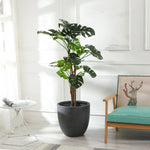 A large contemporary planter stands next to a small round table with a glass of water and magazine, and a wooden chair with a deer cushion, all in a bright room with white walls.