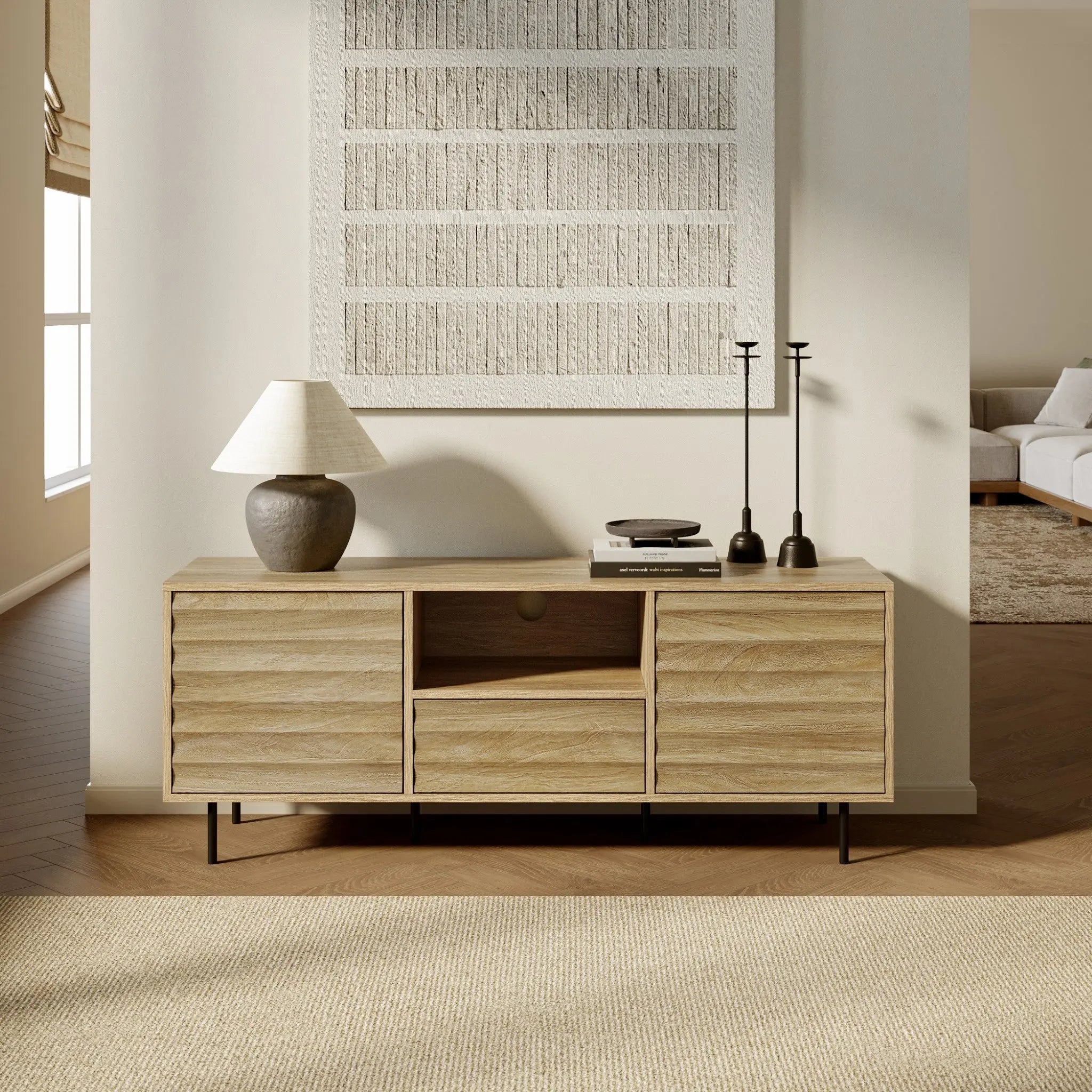 Light walnut-colored wood defines this wooden sideboard, topped with a lamp, books, and candlesticks. A textured wall art piece hangs above, adding to the mid-century modern charm. The room is bright with natural light and offers a glimpse of the adjoining living space.