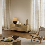 A modern living room features a mid-century modern design with a light walnut-colored wood sideboard, table, and chair. A lamp and decor items adorn the sideboard, which includes a cable management hole. Sheer curtains cover tall windows, complementing the elegant wooden flooring.