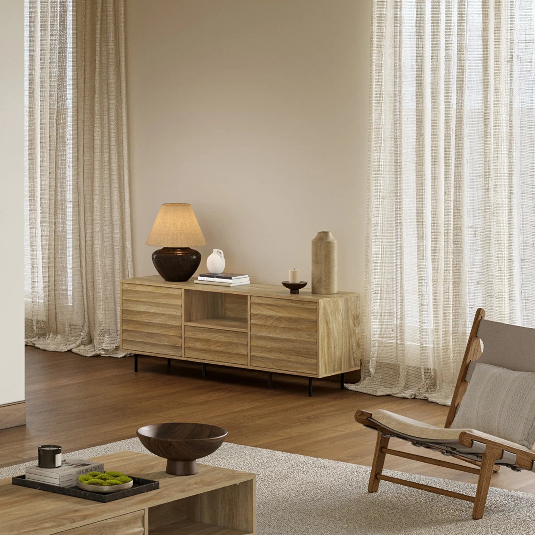 A modern living room features a mid-century modern design with a light walnut-colored wood sideboard, table, and chair. A lamp and decor items adorn the sideboard, which includes a cable management hole. Sheer curtains cover tall windows, complementing the elegant wooden flooring.