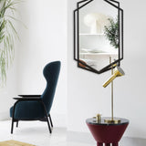 A modern living space with a black armchair, a gold lamp on a maroon round table, a hexagon shape black wall mirror, and a large green plant.