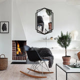 Modern living room with a black rocking chair, small potted tree, fireplace, and a black wall mirror in a hexagon shape reflecting part of the modern style space.