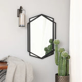A black wall mirror in a hexagon shape reflects a potted cactus beneath it, next to a modern wall sconce and a gray throw on a wooden bench.