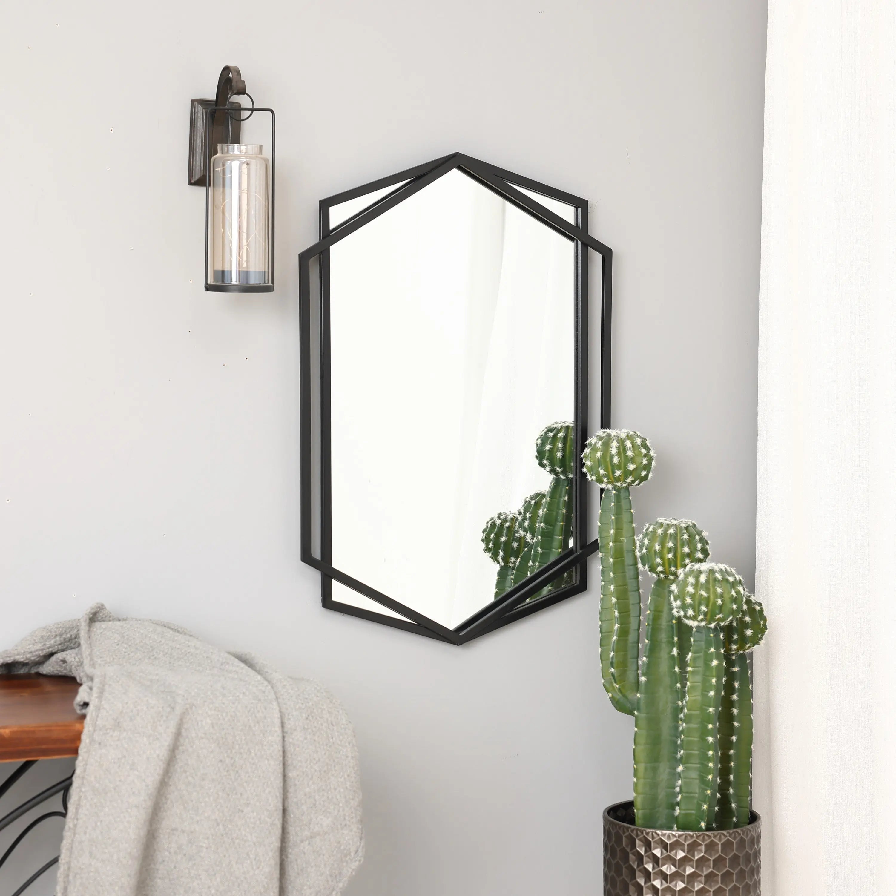 A black wall mirror in a hexagon shape reflects a potted cactus beneath it, next to a modern wall sconce and a gray throw on a wooden bench.