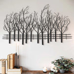Contemporary style wall decoration featuring metal wall art of leafless trees above a shelf with candles, books, and a potted plant on a light-colored wall adds elegant nature theme wall decor to any space.