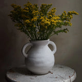 A vintage-inspired white ceramic vase with two handles, filled with yellow flowers, graces a weathered round table against a plain background.