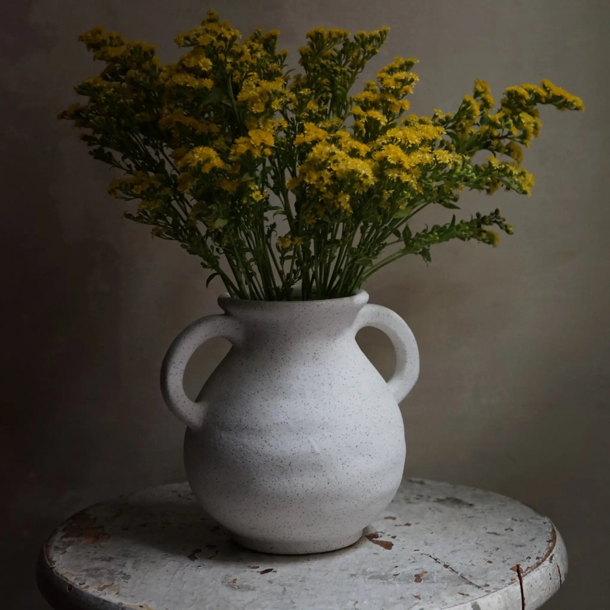 A vintage-inspired white ceramic vase with two handles, filled with yellow flowers, graces a weathered round table against a plain background.