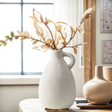A vintage speckled white vase with a handle holds dried golden branches on a wooden table, alongside a smaller beige terracotta vase, enhancing the homes decor in front of a window.