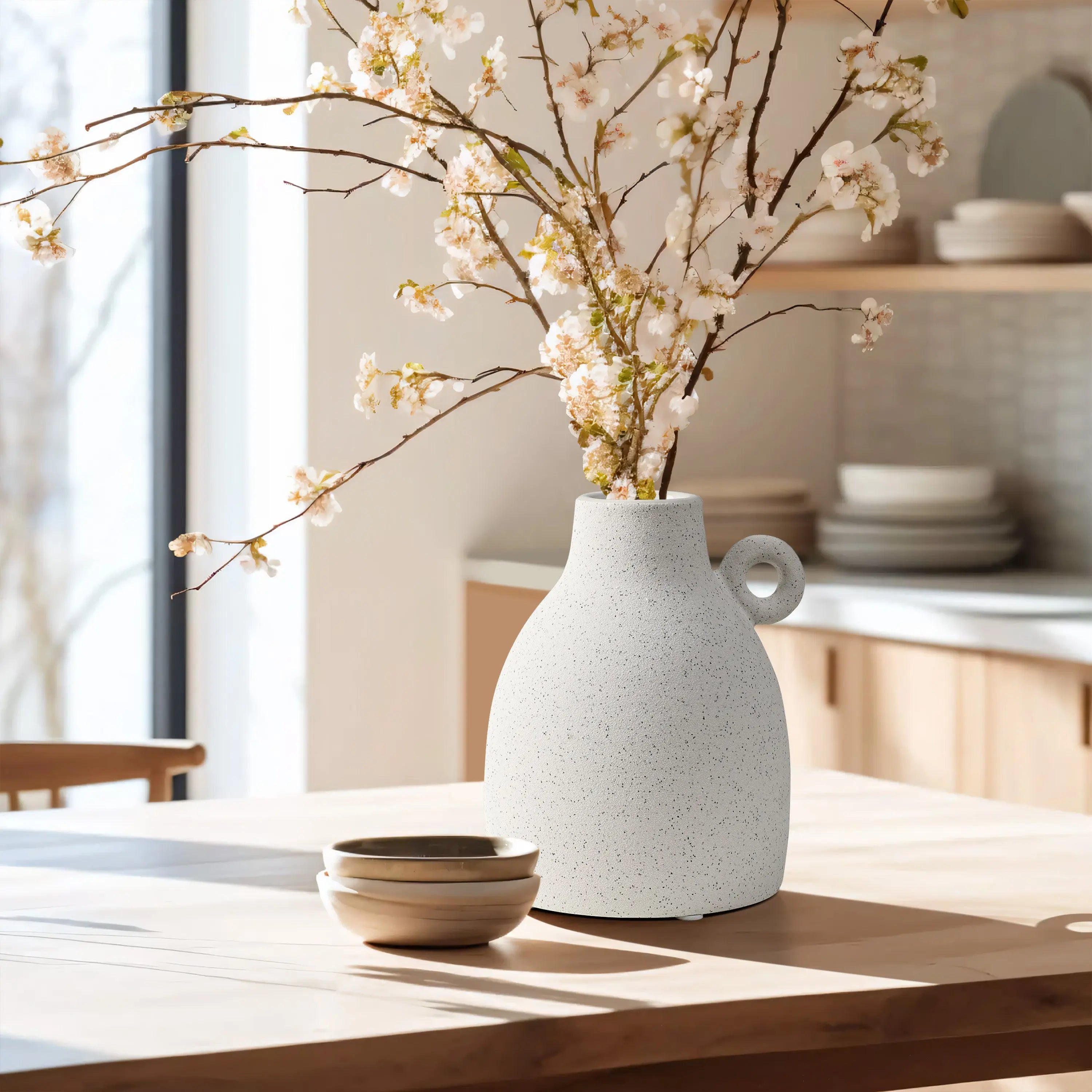A vintage-inspired, white speckled vase holds cherry blossom branches on a wooden table in a sunlit kitchen.