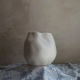 A textured ceramic vase in ivory white, with an uneven shape, sits elegantly as an accent piece on a floral-patterned cloth against a neutral, textured wall.