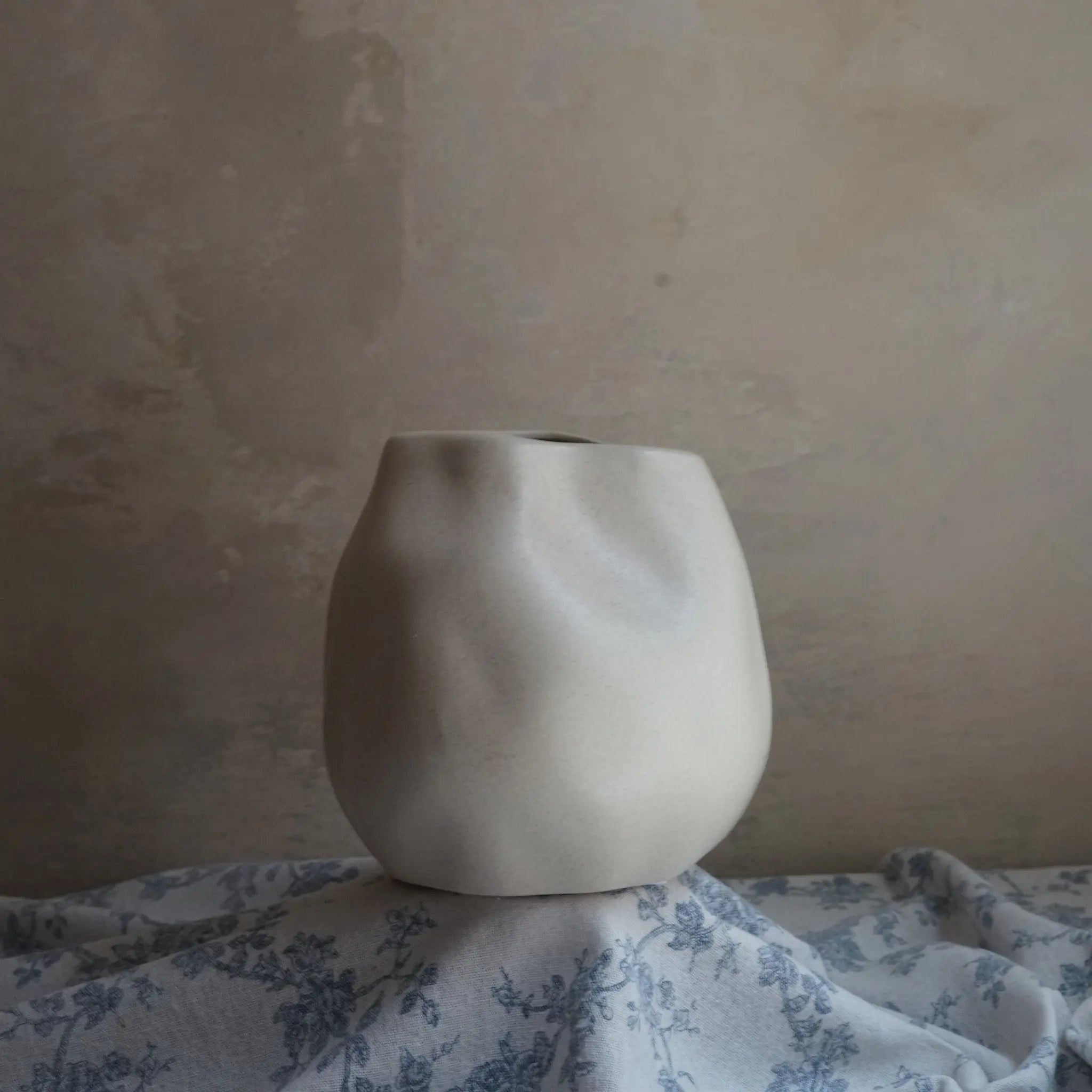 A textured ceramic vase in ivory white, with an uneven shape, sits elegantly as an accent piece on a floral-patterned cloth against a neutral, textured wall.