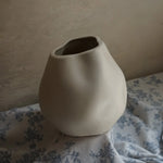 An ivory white misshapen ceramic vase serves as an accent piece, sitting elegantly on blue and white floral fabric against a plain wall.