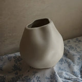 An ivory white misshapen ceramic vase serves as an accent piece, sitting elegantly on blue and white floral fabric against a plain wall.