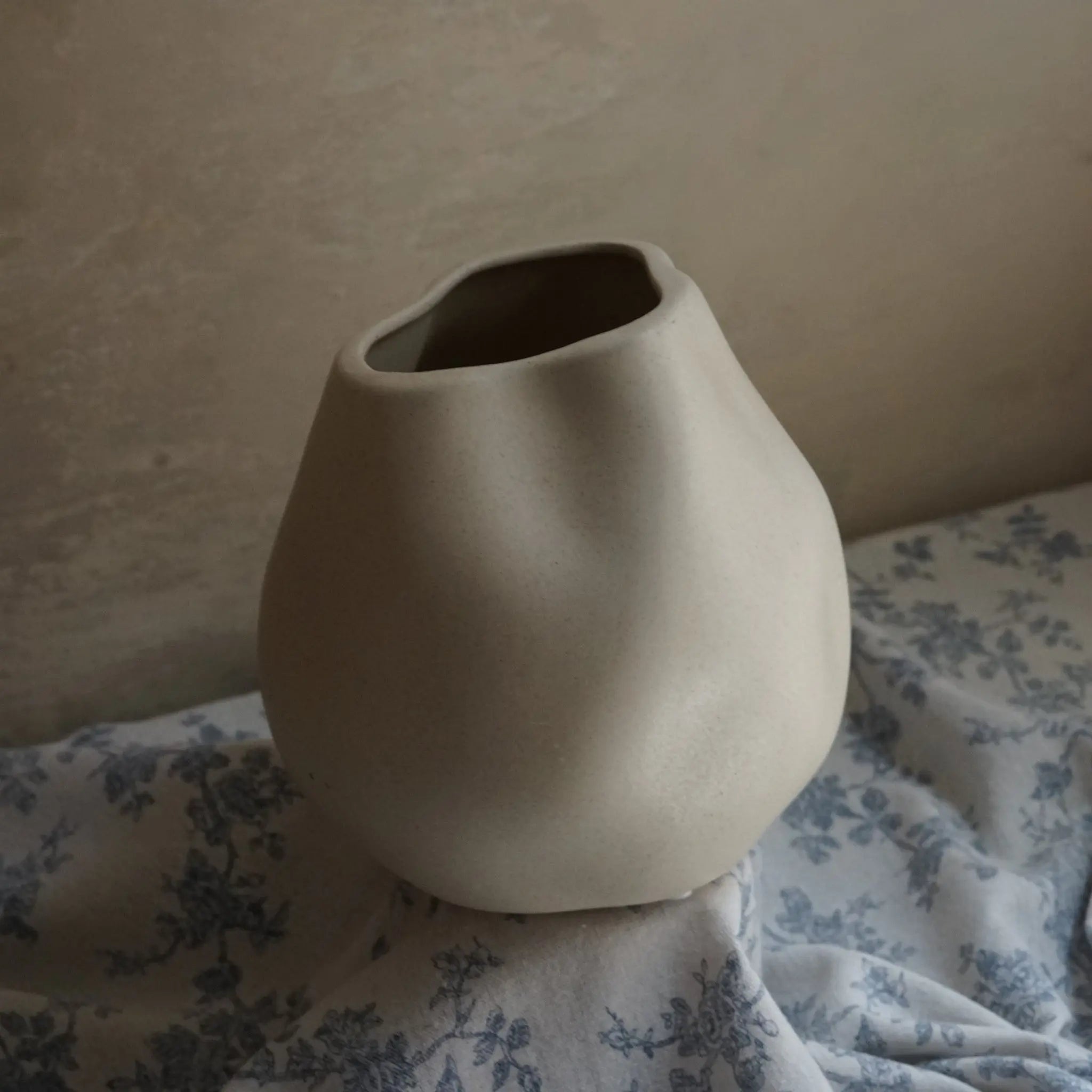 An ivory white misshapen ceramic vase serves as an accent piece, sitting elegantly on blue and white floral fabric against a plain wall.