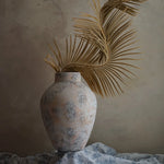 A textured ceramic vase with dried palm leaves, set against a white marble-textured background and draped fabric, creates an elegant home decor statement.