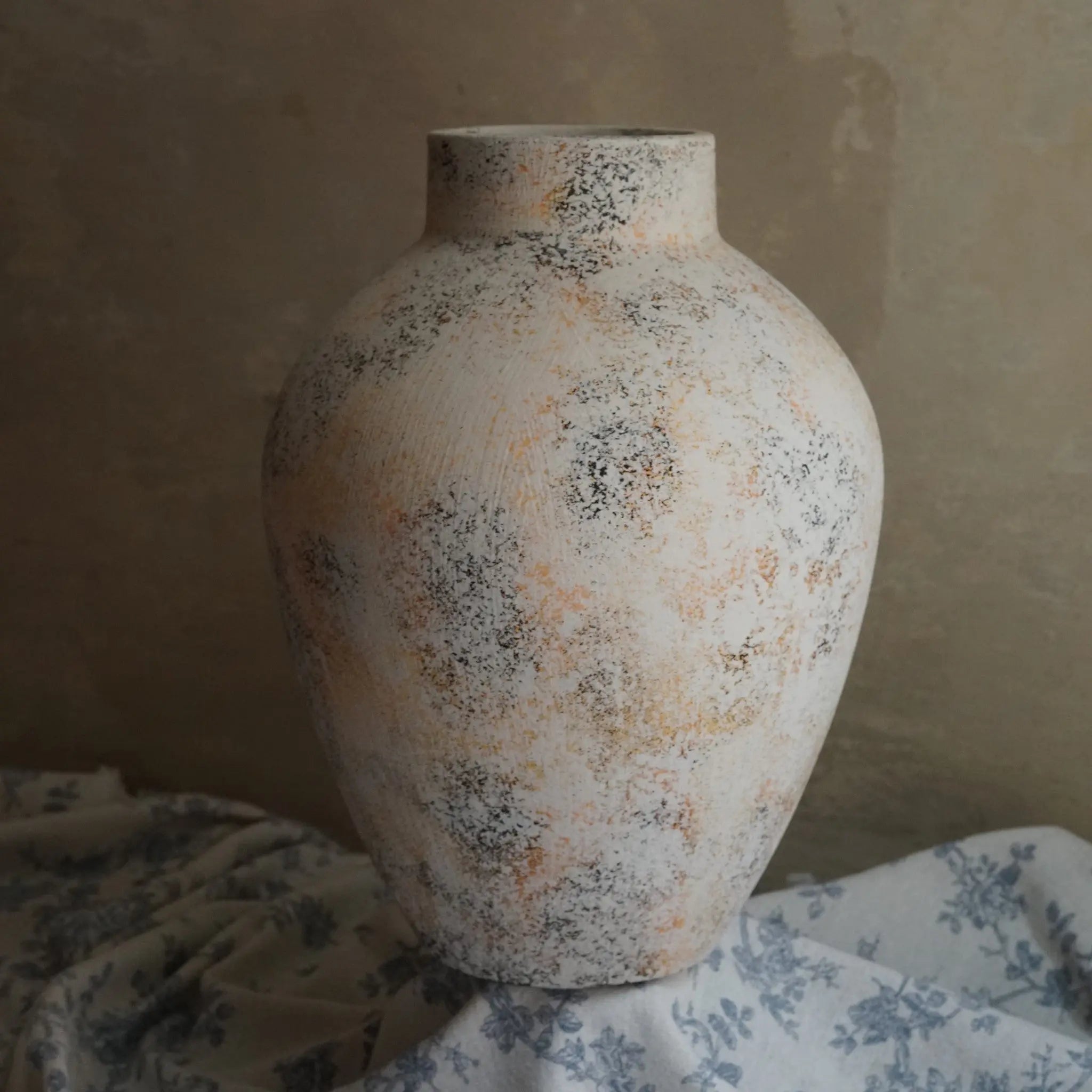 A round, textured ceramic vase with a speckled white marble texture of gray and orange pattern sits on draped floral fabric against a muted background, making it an exquisite piece of home decor.