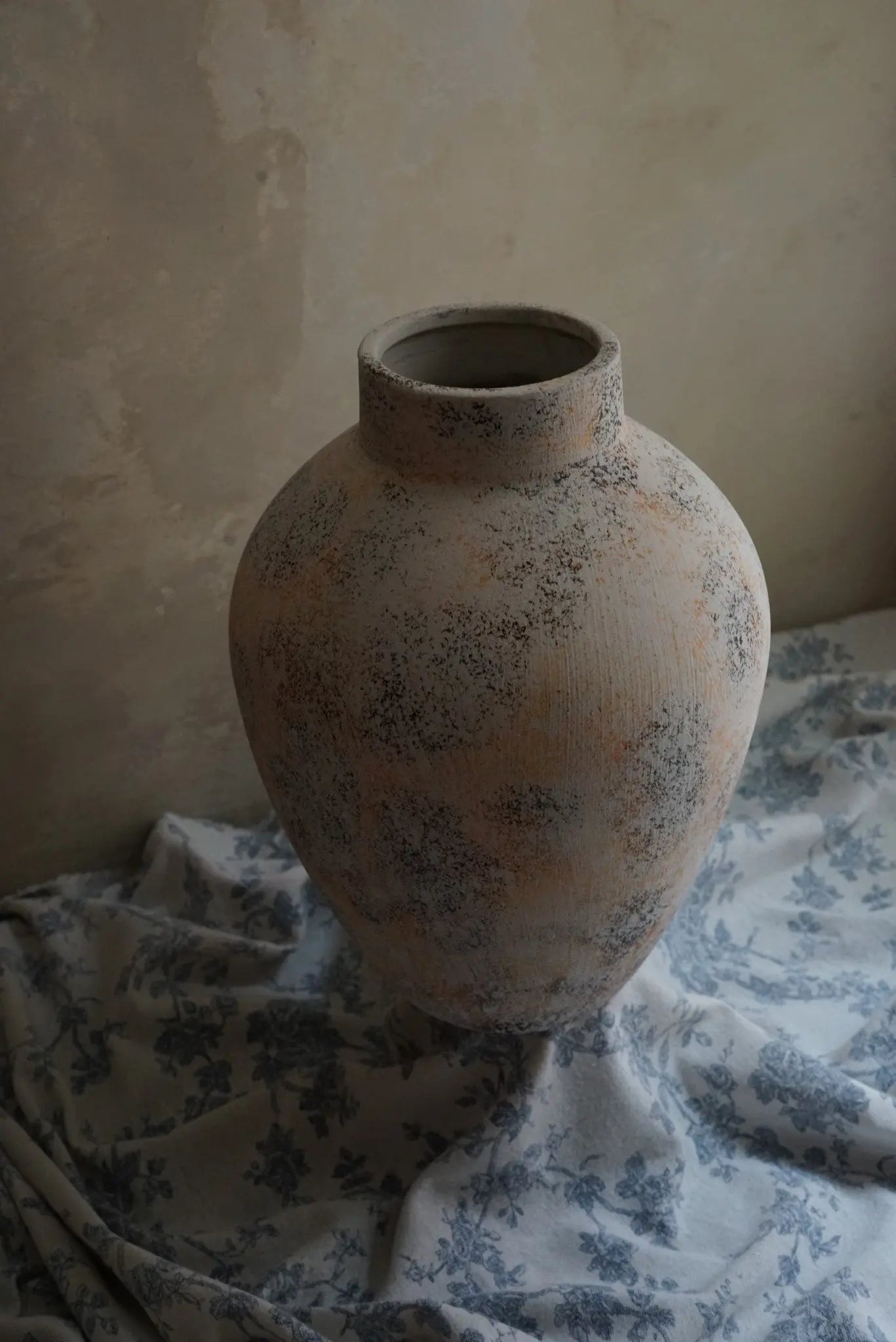 A large, textured ceramic vase enhances the floral-patterned fabric backdrop against a light wall with a subtle white marble texture, creating an elegant home decor statement.
