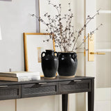 Two black ceramic vases with a delicate black glaze and branches rest on a black table with drawers. A stack of books and a framed picture lean against a cream-colored wall under a gold door handle, creating an elegant contrast.