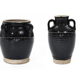 Two black ceramic jugs with handles, showcasing a glossy black glaze. The left jug is 9.84 tall and 7.87 wide, while the right one stands at 10.04 tall and 6.1 wide. Both boast a handcrafted appeal with a beige base that perfectly complements their elegant finish.