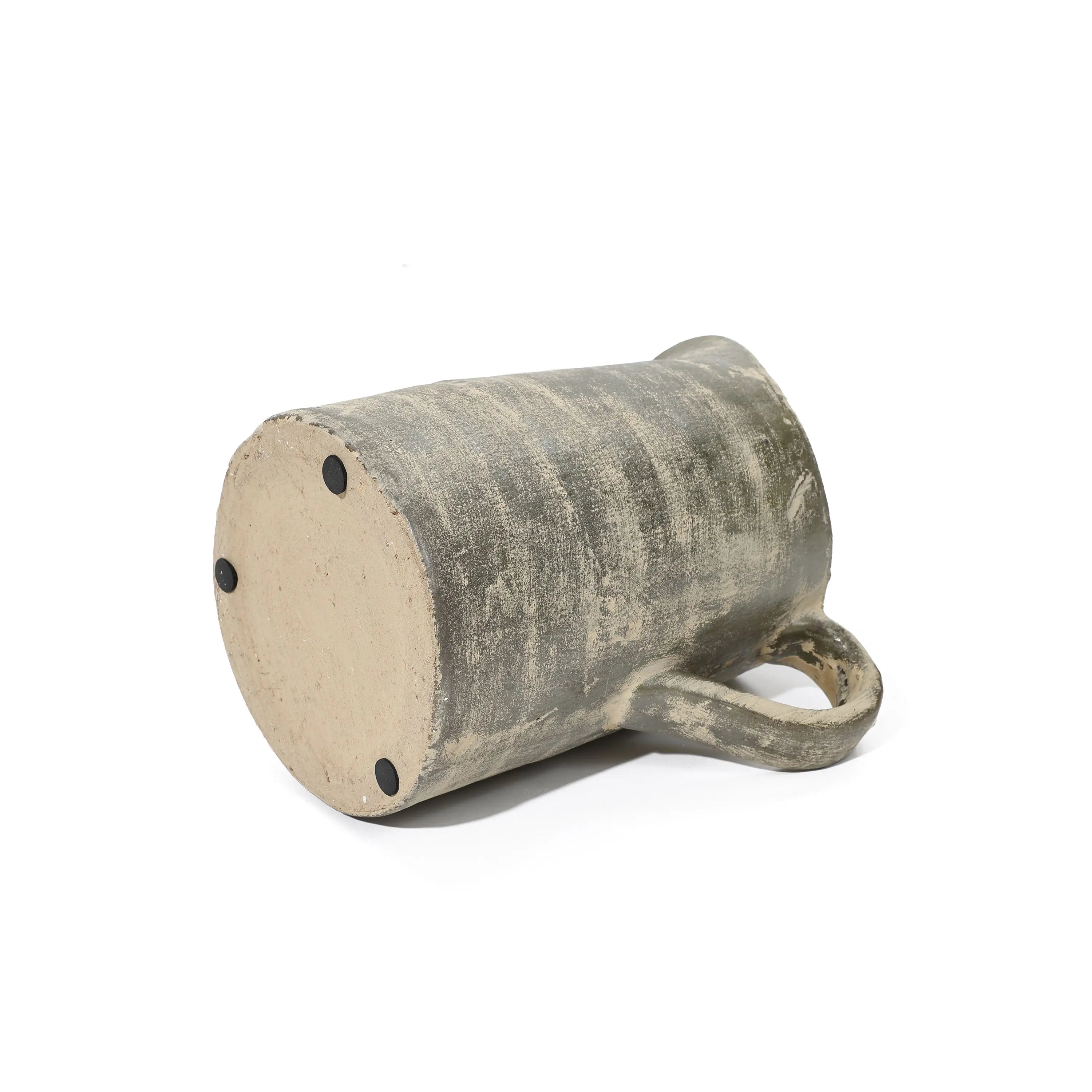 A rustic ceramic mug lies sideways with its handle in view, showcasing artisanal craftsmanship. The mug boasts a textured, earthy terracotta finish and three small pads at the base.