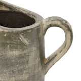 Close-up of a rustic, textured ceramic jug with a round handle and a partially visible open top, showcasing artisanal craftsmanship. The surface appears worn and uneven in color, reminiscent of a terracotta finish.