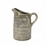An artisanal craftsmanship shines through this rustic, textured ceramic pitcher with a handle, featuring an earthy, muted color and a terracotta finish.