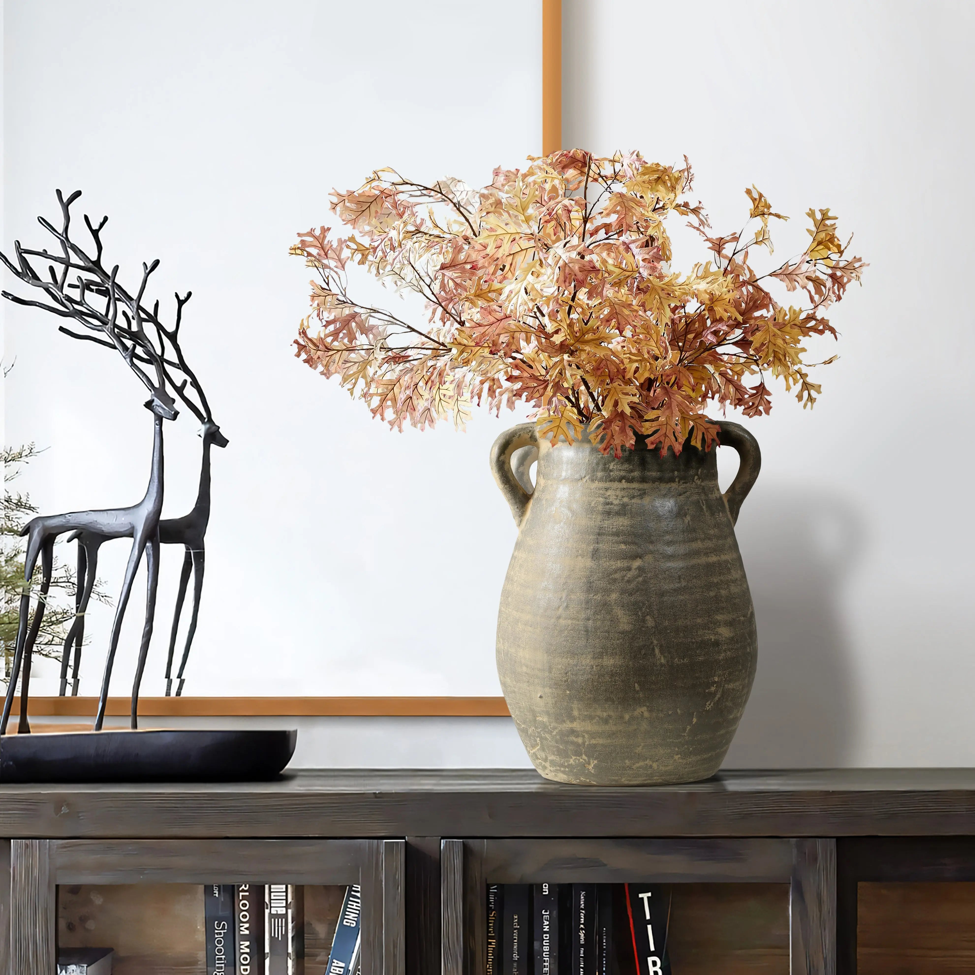 A handcrafted ceramic vase adorned with autumn leaves sits on a wooden shelf, accompanied by a black metal deer sculpture. Below, books add their own historic depth to the scene.