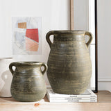 Two large, rustic, brown ceramic pots with handles are displayed on a stack of design books beside a modern abstract painting, radiating historic depth. Each piece resembles a terracotta jug vase, blending timeless craftsmanship with contemporary art.